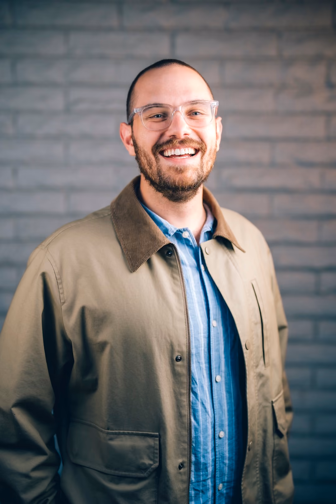 Smiling man with glasses wearing blue shirt and beige jacket standing in front of a gray brick wall.