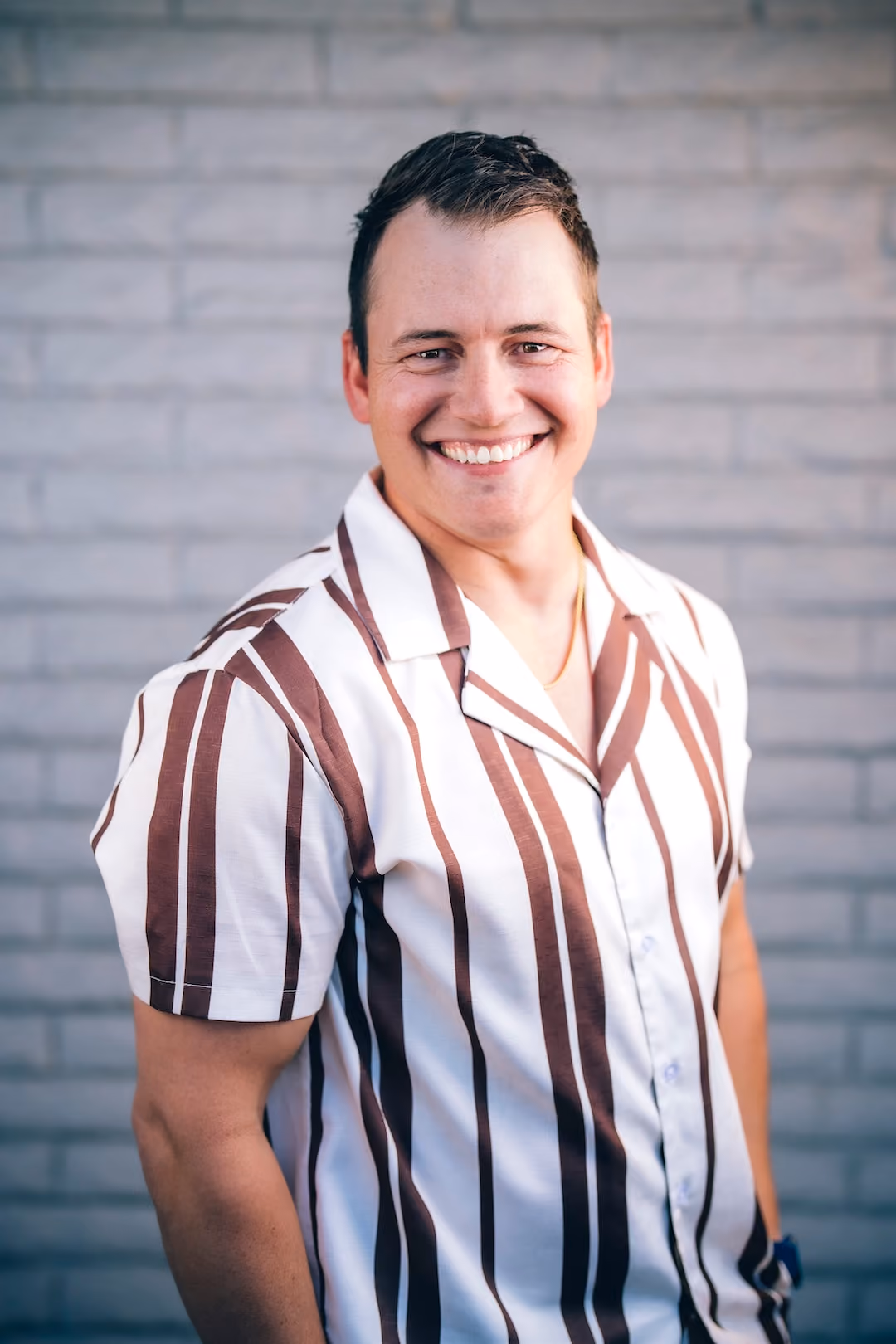 Smiling man in brown and white striped short-sleeve shirt standing in front of a gray brick wall.