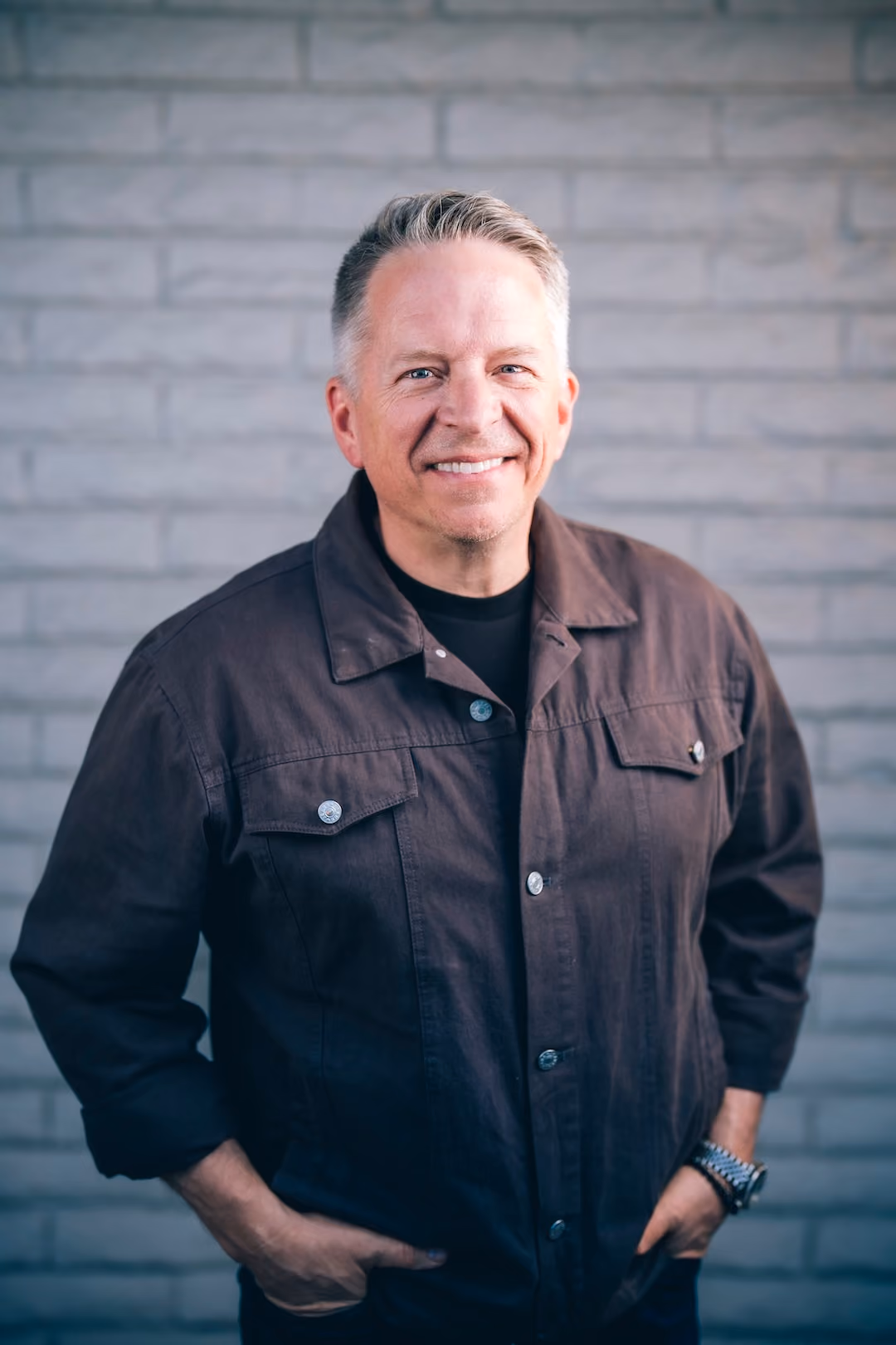 Smiling man with gray hair wearing a dark jacket standing against a light gray brick wall.