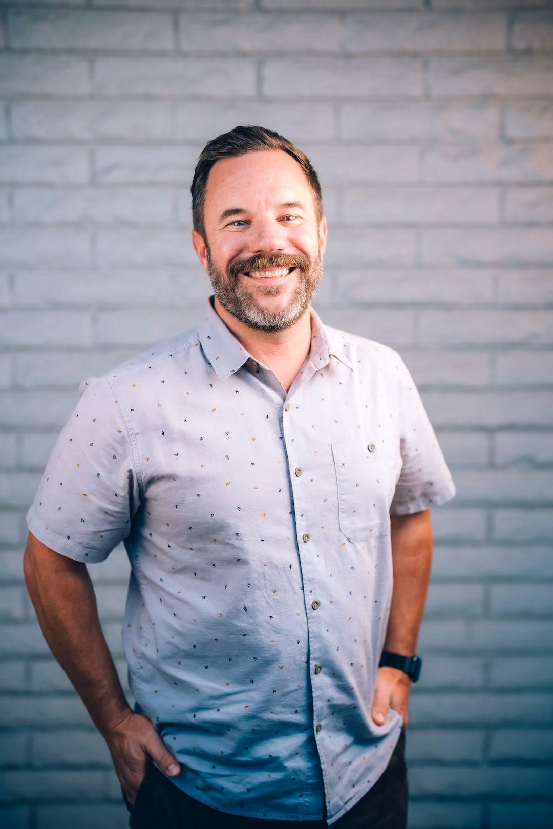 Smiling man with a beard in a light patterned short-sleeve shirt standing before a gray brick wall.