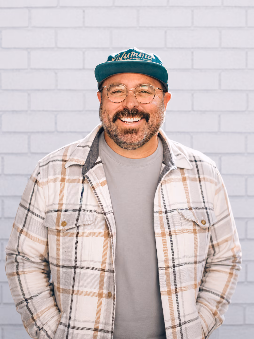 Smiling man with glasses and beard wearing a green cap and plaid shirt against a white brick wall.