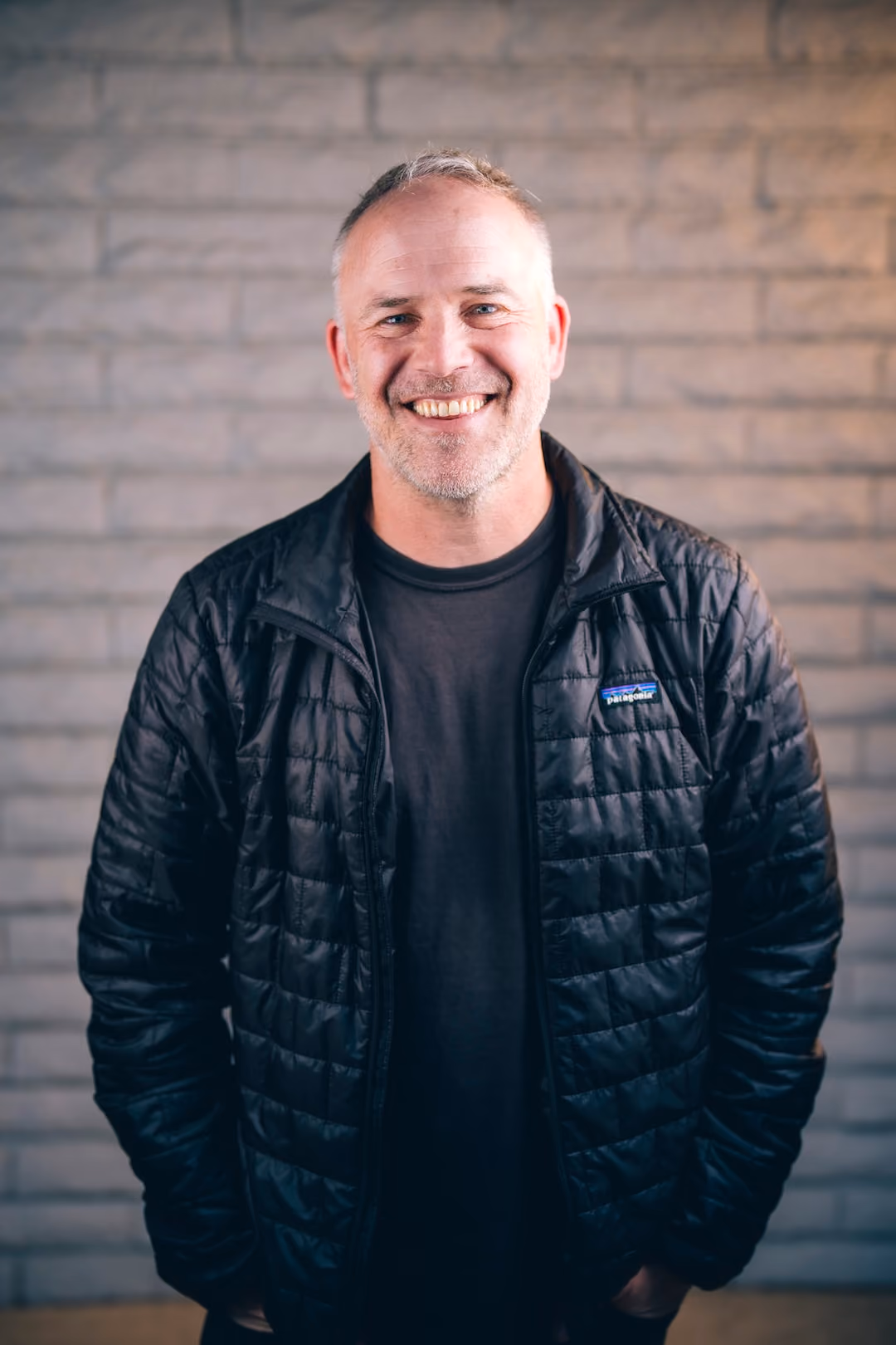 Smiling man with gray hair wearing a black Patagonia jacket stands against a brick wall background.