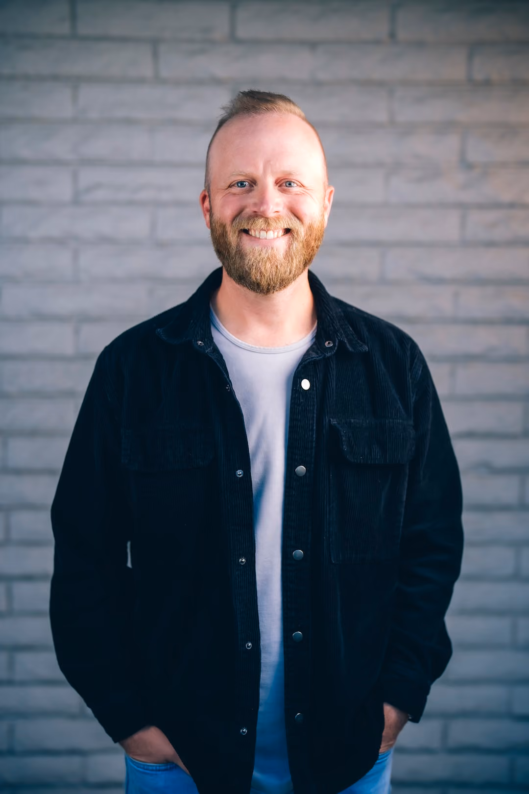 Smiling man with a beard wearing a black jacket and light shirt standing against a brick wall.