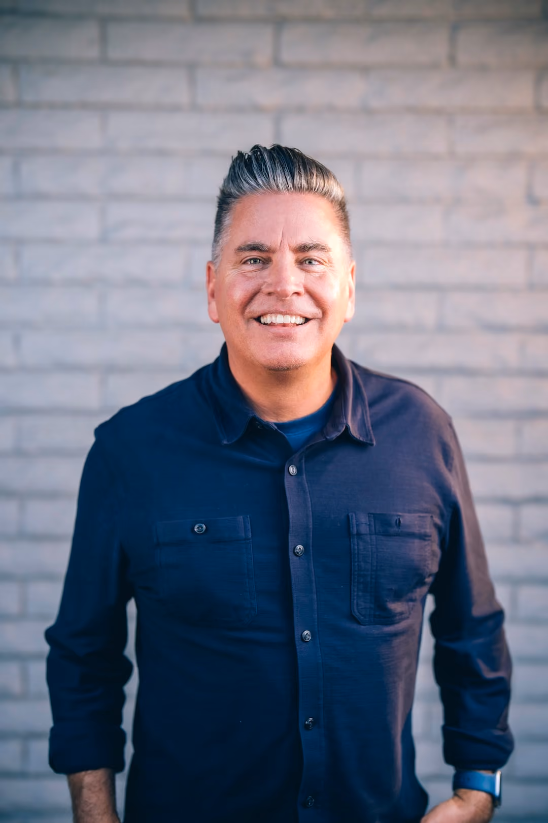 Smiling man with gray slicked-back hair wearing a navy button-up shirt against a brick wall.