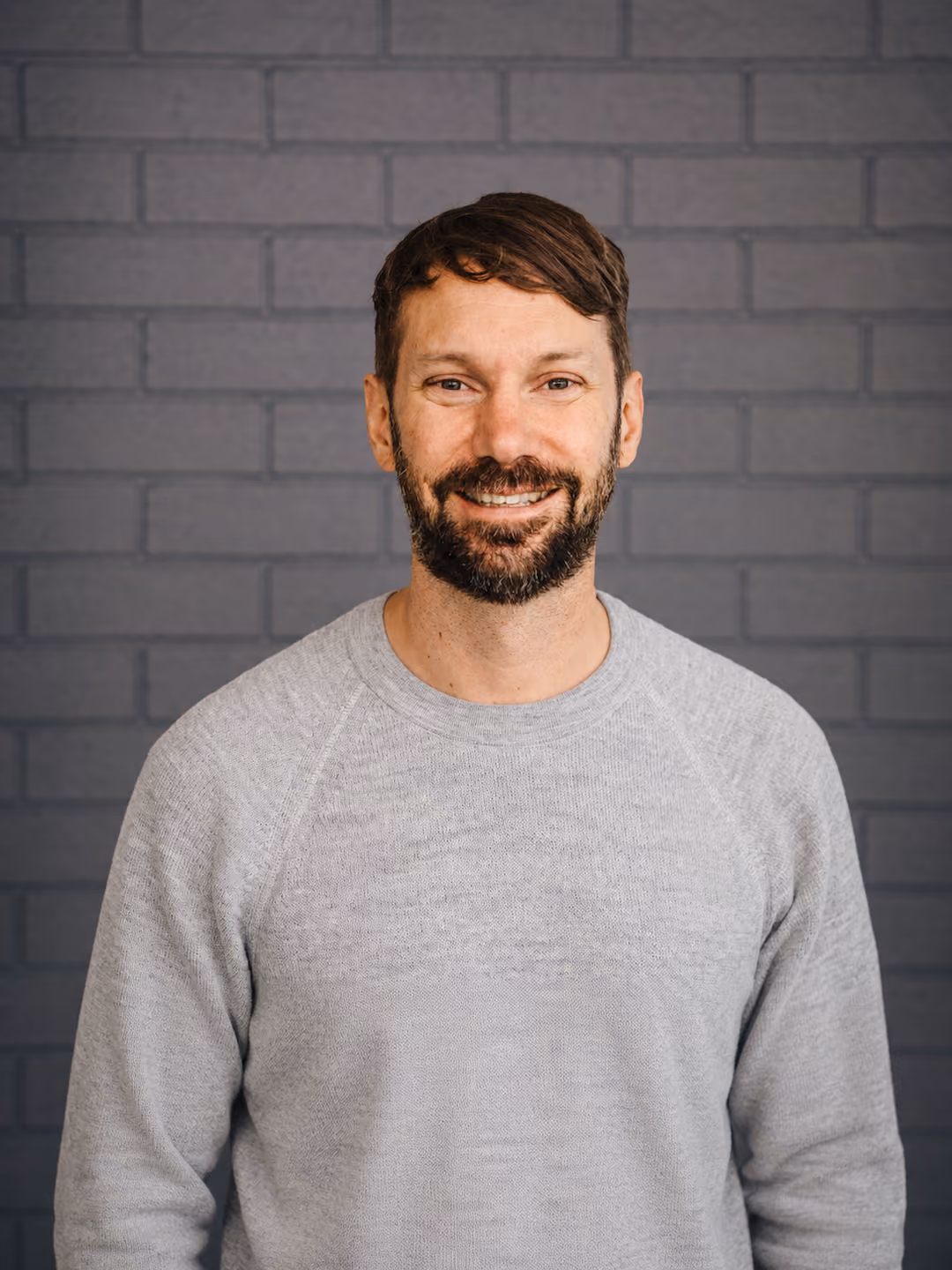 Smiling man with dark hair and beard wearing a gray sweater against a gray brick wall.
