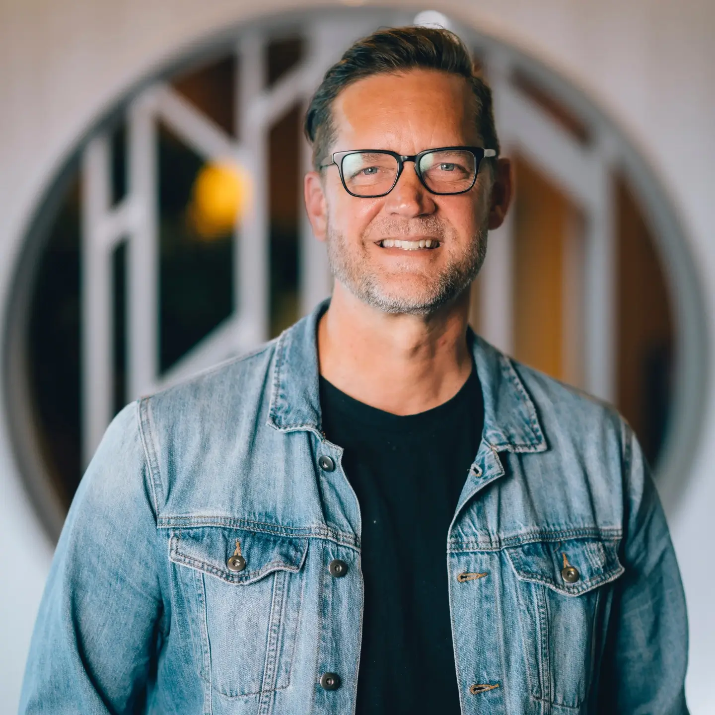 Smiling man with glasses wearing a denim jacket over a black shirt indoors.